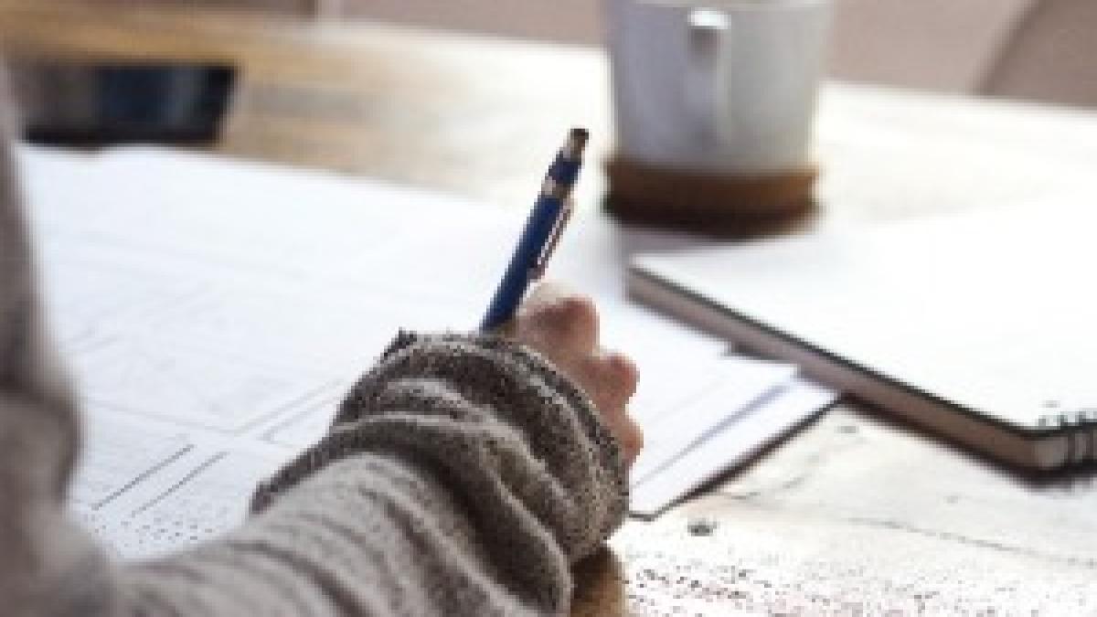 Person writing on brown wooden table near white ceramic mug