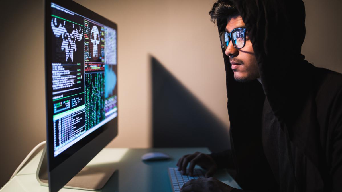 Хакер в капюшоне сидит перед монитором с кодом